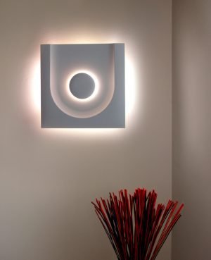 The Cleoni Fundu Wall Light (asymmetric version) features a square design with a circular indentation that extends to the edge on a single side that provides ambient illumination as well as the back of the fitting. Available in gypsum white. Shows the light switched on in a room.